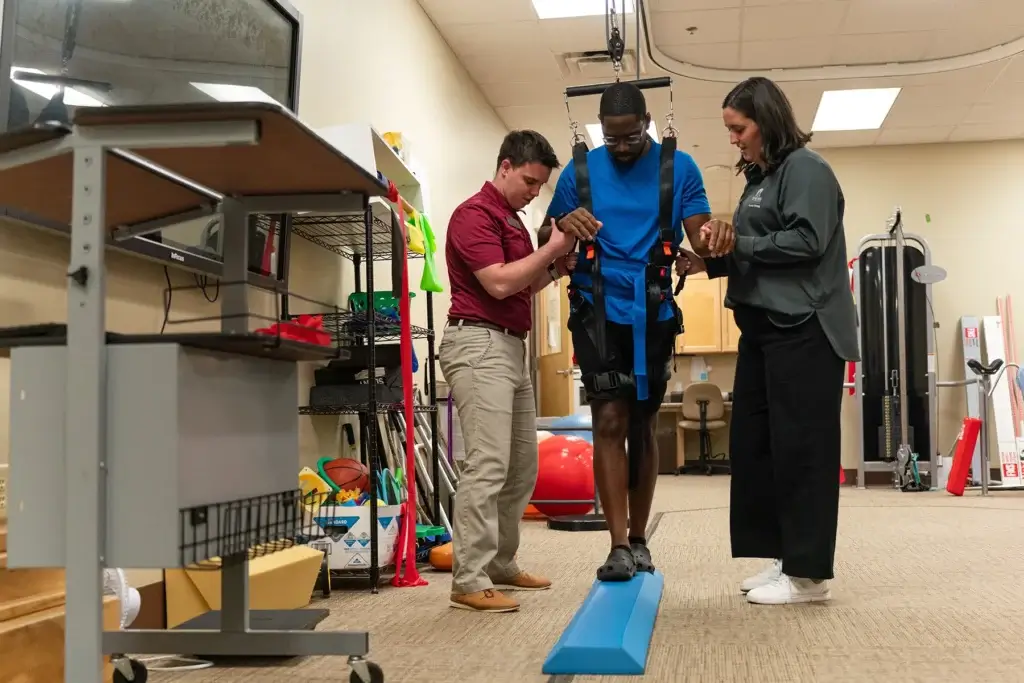 Two physical therapy clinic staff members assist a client to walk on a balance beam