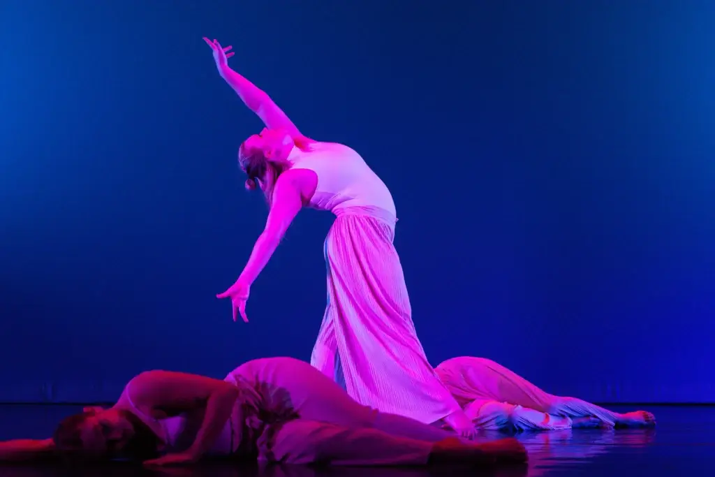 Student dancer leans back and reaches to the sky. Lit pink against a blue background.
